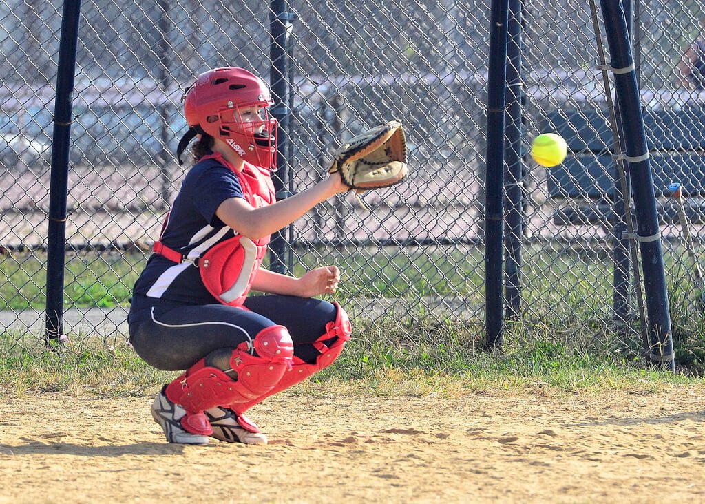 Left-handed fastpitch softball catcher in red gear receiving a pitch with the mitt on her right hand