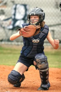 Elite Female fastpitch catcher in dark navy padded chest protector, helmet, and leg guards with leather mitt kneeling on a dirt infield, ready stance