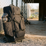 A tall rolling catcher's equipment duffel standing against a chain-link dugout fence